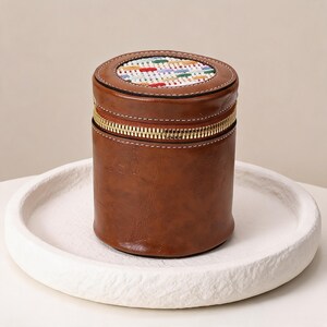 May include: A cylindrical brown leather case with a gold zipper and a colorful woven top. The case is sitting on a white, textured, round tray. The top of the case has a stitched leather border.