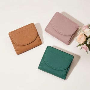 Op de afbeelding: Drie kleine, vierkante portemonnees in bruin, mauve en groen. Elke portemonnee heeft een gebogen klepsluiting en een getextureerde lederen uitstraling. Een klein boeket bloemen staat op de achtergrond.