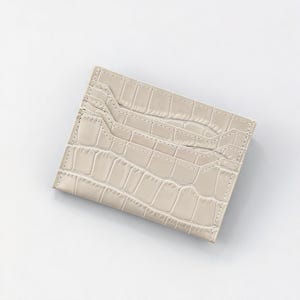 May include: A light beige card holder with a crocodile skin texture. The card holder has multiple slots for cards and a stitched border. The overall design is rectangular and minimalist.