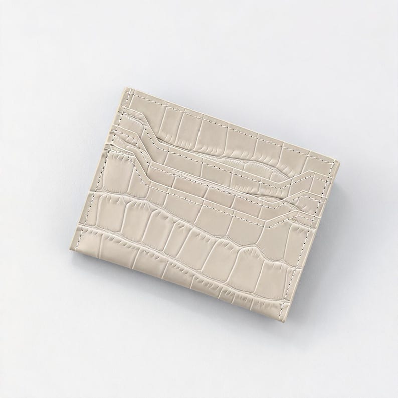 May include: A light beige card holder with a crocodile skin texture. The card holder has multiple slots for cards and a stitched border. The overall design is rectangular and minimalist.