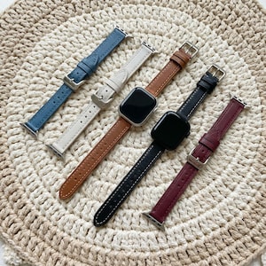 May include: Five watch bands with silver-toned metal buckles and connectors. The bands are in blue, off-white, tan, black, and burgundy. Two of the bands are attached to black smartwatches with rectangular screens and rounded corners.