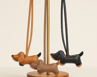 Puede incluir: Tres colgantes de cuero con forma de perro salchicha en marrón, beige y negro, cada uno con una correa a juego. Los colgantes se exhiben en un soporte dorado con una base de madera. Los perros tienen detalles cosidos.