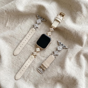 May include: An Apple Watch with a cream-colored leather band and decorative gold and silver accents. The band features a floral design with a gold buckle and silver connectors. The watch face is black.