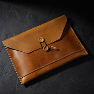 May include: A tan leather document folder featuring a triangular flap closure secured by a black elastic cord looped around two circular brass-colored metal studs. The folder has visible stitching along the edges and a smooth, matte finish.