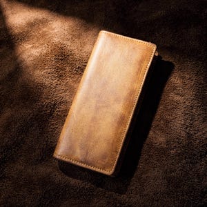 May include: A rectangular, tan leather wallet with a distressed finish. The wallet is stitched around the edges and appears to be made of high-quality leather. The wallet is lying on a brown, textured surface.