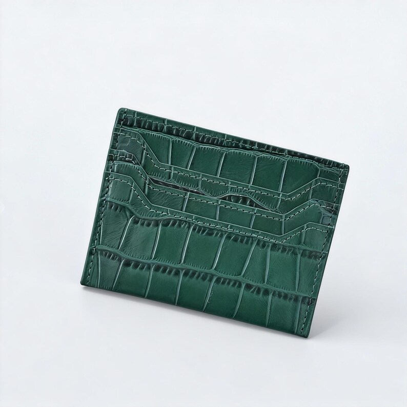 May include: A dark green card holder with a crocodile skin texture. The rectangular wallet has multiple card slots and white stitching around the edges. The wallet is made of a durable material.
