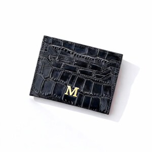 May include: Black card holder with a crocodile texture and a gold letter "M" on the front. The card holder has multiple slots for cards and a sleek, modern design. The card holder is made of a durable material.