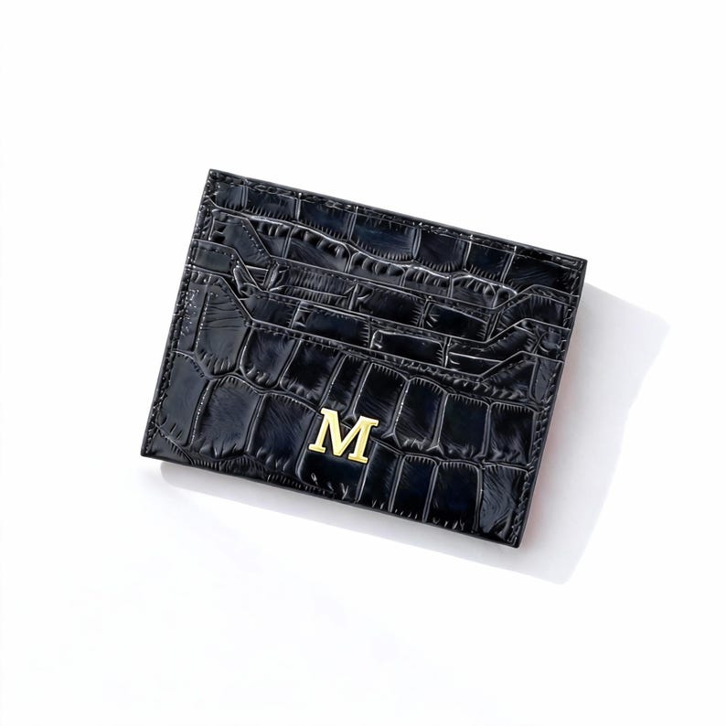May include: Black card holder with a crocodile texture and a gold letter "M" on the front. The card holder has multiple slots for cards and a sleek, modern design. The card holder is made of a durable material.