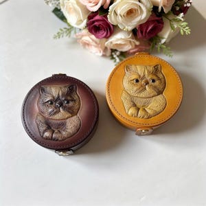 May include: Two round leather cases, one brown and one tan, each featuring a detailed embossed image of a cat. The cases have a clasp closure and are likely designed for small items. A bouquet of flowers is in the background.