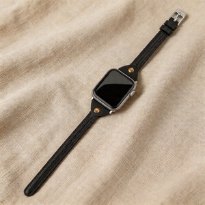 May include: A black leather watch band with a silver buckle and a silver watch face. The band is attached to the watch face with brass-colored rivets. The watch is displayed on a beige fabric surface.