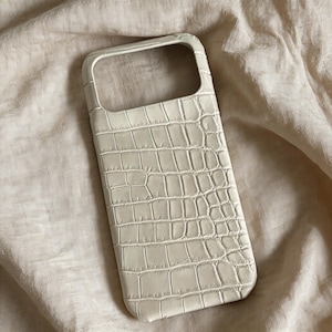 Puede incluir: Funda de teléfono de color crema con textura de piel de cocodrilo. La funda tiene forma rectangular con esquinas redondeadas y un recorte para la cámara. Fabricada con un material resistente, protege el teléfono.