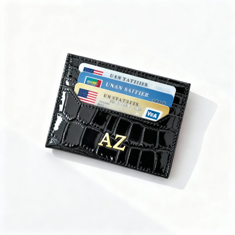 May include: A black, glossy cardholder with a crocodile skin texture. The cardholder has the gold letters "AZ" on the front and holds three cards with American flag designs. The cardholder is designed to hold credit cards and identification.