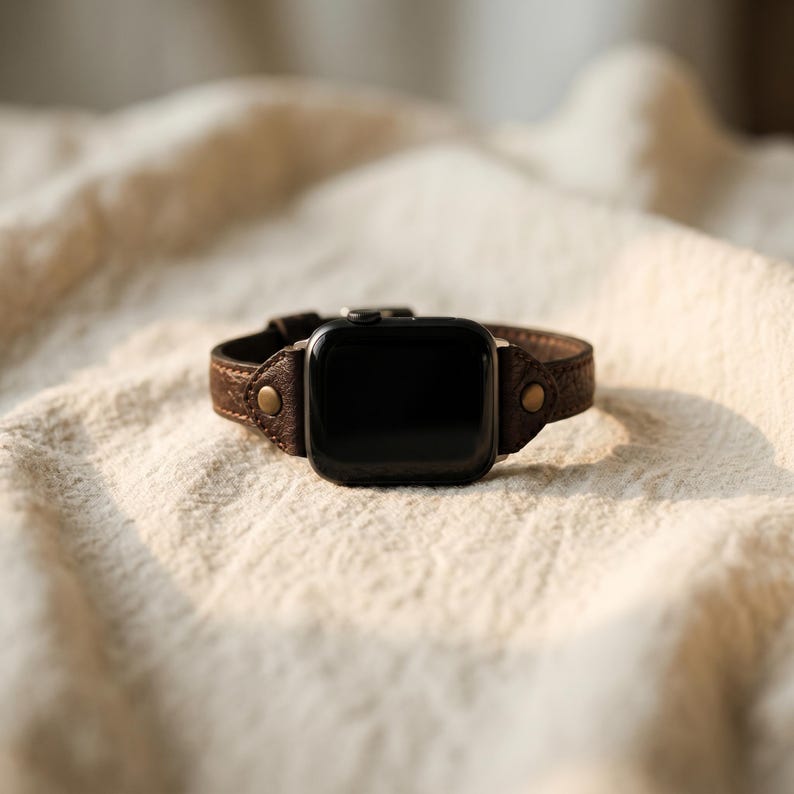 May include: A brown leather watch band with a black watch face. The band is made of textured leather and has two small, round, brass-colored rivets on each side of the watch face. The watch is resting on a cream-colored, textured fabric.
