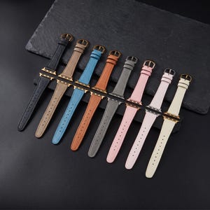May include: A collection of eight leather watch bands for smartwatches, arranged in a diagonal row. The bands feature gold-toned metal buckles and connectors, and come in black, tan, blue, orange, gray, light pink, pale pink, and cream colors.