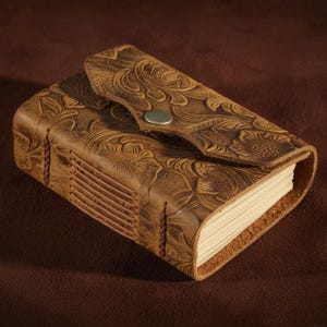 May include: A brown leather journal with a floral embossed design. The journal has a flap closure with a metal snap and stitched binding. The pages are visible and appear to be blank.