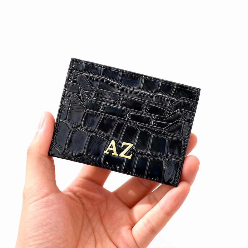 May include: Black card holder with a crocodile texture and the gold letters "AZ". The card holder has multiple slots for cards. The card holder is held in a hand.