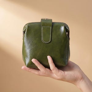 Puede incluir: Un pequeño monedero de cuero verde oliva con cierre de broche. El monedero se sostiene en una mano, mostrando su tamaño compacto y textura suave. El cierre se asegura con un botón metálico. Aproximadamente 10 cm de ancho.
