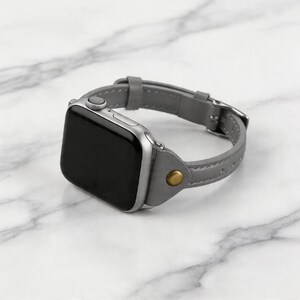 May include: A silver smartwatch with a gray leather band. The band features a buckle closure and a gold-colored rivet. The watch face is black and the watch is displayed on a white marble surface.