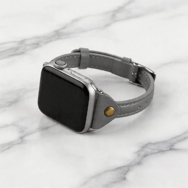 May include: A silver smartwatch with a gray leather band. The band features a buckle closure and a gold-colored rivet. The watch face is black and the watch is displayed on a white marble surface.