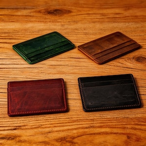 May include: Four leather cardholders in green, brown, burgundy, and black are displayed on a wooden surface. Each cardholder has a rectangular shape with multiple card slots and stitched edges. The leather has a distressed, textured appearance.
