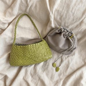 Könnte beinhalten: Eine hellgrüne, gewebte Handtasche mit geflochtenem Henkel liegt neben einem grauen Beutel mit Kordelzug. Die Handtasche hat eine rechteckige Form mit einem Webmuster. Der Beutel hat einen gelben Akzent am Rand und grüne Akzente an den Kordelenden.