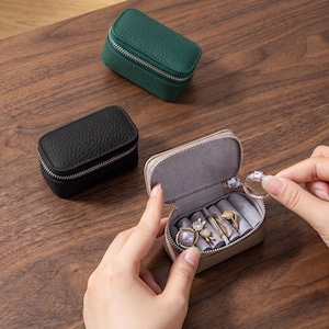 May include: Three small rectangular jewellery cases with textured surfaces in green, black, and beige. The beige case is open, revealing a grey velvet interior with slots for rings and small accessories, while a hand places a silver ring inside.
