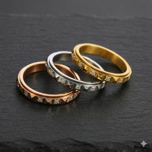 May include: Three rings in rose gold, silver, and gold, each with a band of small, square-cut gemstones. The rings are displayed on a dark, textured surface, showcasing their metallic shine and intricate design. The rings are a classic jewelry item.
