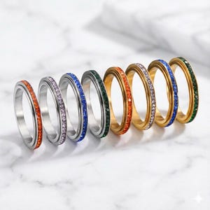 May include: A collection of seven rings with colorful gemstones. The rings feature a variety of colors, including orange, purple, blue, and green. Some rings have a silver finish, while others are gold.
