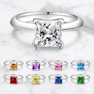 May include: A collection of silver rings, each featuring a square-cut gemstone. The top ring has a clear stone, while the others display various colors: orange, purple, pink, blue, red, yellow, dark blue, and green. The rings are displayed on a white surface.