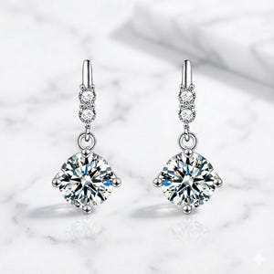May include: A pair of silver-toned drop earrings featuring a round, faceted clear stone suspended from a vertical bar. The bar is accented with two smaller, round, faceted clear stones set in a row above the larger dangling circular stone.