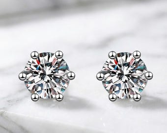 May include: A pair of sparkling, clear diamond stud earrings. Each earring features a large, faceted gemstone held in place by six small, silver-colored prongs. The earrings are set against a white and gray marble background.