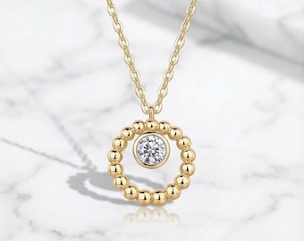 Può includere: Una collana color oro con un ciondolo. Il ciondolo presenta una gemma trasparente al centro, circondata da un anello di piccole perle color oro. La catena è anch'essa color oro e ha un design delicato. La collana è mostrata su uno sfondo di marmo bianco.