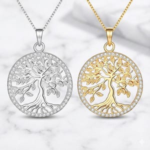 May include: Two tree of life pendant necklaces. One is silver-toned, the other gold-toned. Both feature a circular design with a tree silhouette and a border of small, clear stones. The pendants hang from delicate chains.