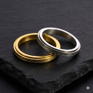 May include: Two rings, one gold-toned and one silver-toned, are interlocked on a dark surface. Both rings have a textured band design. The gold ring is on the bottom, with the silver ring resting on top.