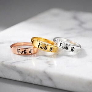 May include: Three metal rings in rose gold, yellow gold, and silver tones. Each ring features a flat, polished band engraved with the black cursive text "Fuck it" centered on the front surface, showcasing a simple and clean design.
