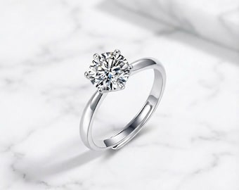 May include: A silver ring with a large, round, clear gemstone. The gemstone is set with six prongs. The ring band is thin and adjustable. The ring is displayed on a white marble surface.