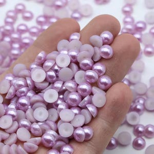 Peut inclure: Une collection de petites perles artificielles demi-rondes de couleur violet clair avec une finition lisse et brillante. Chaque pièce présente une base plate, blanche et circulaire sur le dessous, conçue pour une fixation facile sur des surfaces.
