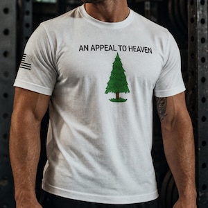 May include: A white short-sleeved t-shirt featuring the black text AN APPEAL TO HEAVEN above a green pine tree graphic. The right sleeve displays a small black and white American flag patch. The design is centred on the chest of the garment.