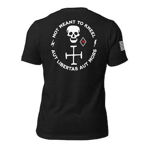 May include: Black t-shirt with a white graphic on the back. The graphic includes a skull, a cross, lightning bolts, and the text "NOT MEANT TO KNEEL AUT LIBERTAS AUT MORS". A small American flag design is on the right sleeve.