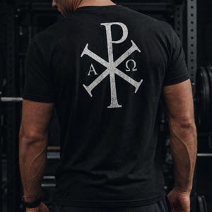 May include: A black short-sleeved t-shirt featuring a large, distressed white Chi-Rho symbol printed on the back. The design includes the Greek letters Alpha and Omega positioned on the left and right sides of the central cross-like emblem.