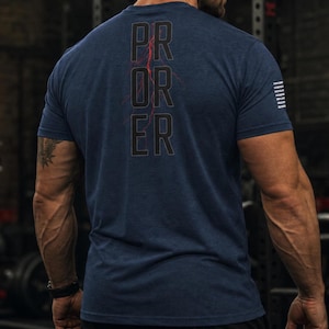Pode incluir: Uma t-shirt azul-marinho de manga curta com o texto PR OR ER empilhado verticalmente nas costas. Um gráfico de um relâmpago vermelho atravessa o centro das letras. Um pequeno gráfico de uma bandeira branca está impresso na manga direita.