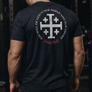 May include: A dark navy blue short-sleeve t-shirt featuring a white Jerusalem cross graphic on the back. The design includes the circular text THE WILL OF GOD IS OUR SHIELD AND OUR SWORD and the red text DEUS VULT positioned below the cross.