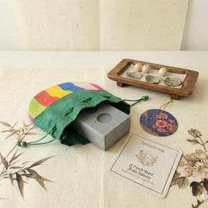 May include: A green drawstring bag with a colorful striped top holds a gray concrete block with a circular indentation. A small wooden tray with stones and spheres sits nearby, along with a floral tag and a card with text.