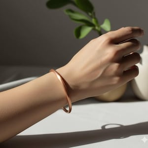 Peut inclure: Un bracelet jonc en cuivre massif lisse avec un profil arrondi. Le bracelet présente un design circulaire continu et sans couture avec une finition métallique polie qui reflète la lumière, créant des reflets subtils sur sa surface courbe.