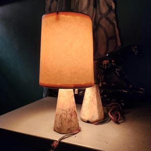 May include: A table lamp with a cylindrical, warm-toned fabric shade and a conical base featuring a marbled, cream and reddish-brown pattern. A second, matching lamp base sits partially visible behind it, both with frayed fabric cords attached.