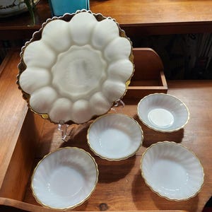 May include: A white, scalloped serving platter with a gold rim, accompanied by three small matching bowls. The platter has a unique design, and the bowls have a similar gold trim. The set is displayed on a wooden surface.