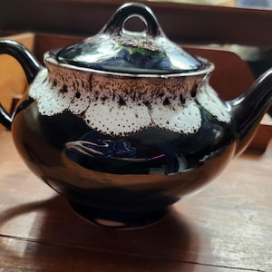 May include: A glossy black ceramic teapot featuring a rounded body, a curved handle, and a spout. The upper portion of the teapot and the matching lid are decorated with a textured, white drip-glaze pattern that contrasts with the dark base.