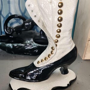 May include: A decorative ceramic boot vase in black and white with gold-colored button accents. The boot has a high heel and sits on a matching base. The vase is a unique home decor item.