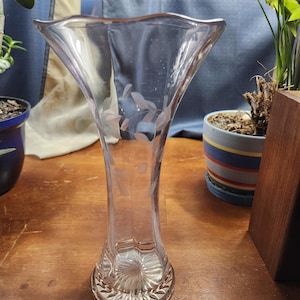 May include: A clear glass vase with a flared top and etched leaf design. The vase has a slender neck and a decorative base. The vase is sitting on a wooden surface.