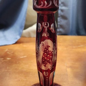 May include: A ruby red glass candlestick holder with etched white floral and geometric designs. The candlestick has a wide base, a slender stem, and a flared top. The etched designs include swirls, dots, and a stylized leaf pattern.